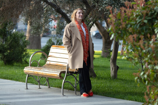 a woman tanding near the bench with metal armrests in an outdoor setting. She is smiling warmly, resting her chin on her hand, and wearing a tan coat over a white shirt and orange scarf
