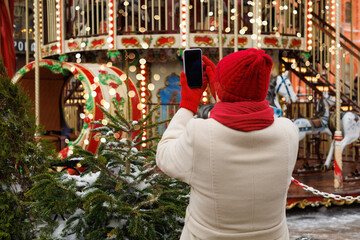Fototapeta premium Senior woman in a warm coat and bright red accessories take photo of colorful, beautifully illuminated carnival Christmas carousel. Diversity, happy and elderly people in retirement.