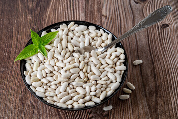 White beans in a black bowl over brown wooden background. Raw organic kidney beans or haricot prepared for cooking vegetarian baked beans. Harvesting, plant protein.