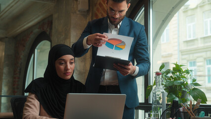 Muslim woman in hijab female businesswoman working laptop at office desk Arabian Indian businessman male business man boss come analyze paperwork documents check results smile approve good job praise
