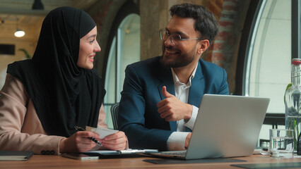Happy business partners Arabian couple colleagues male female count bills taxes online payment bank on laptop smiling man muslim woman hijab businessman businesswoman thumb up gesture good job office