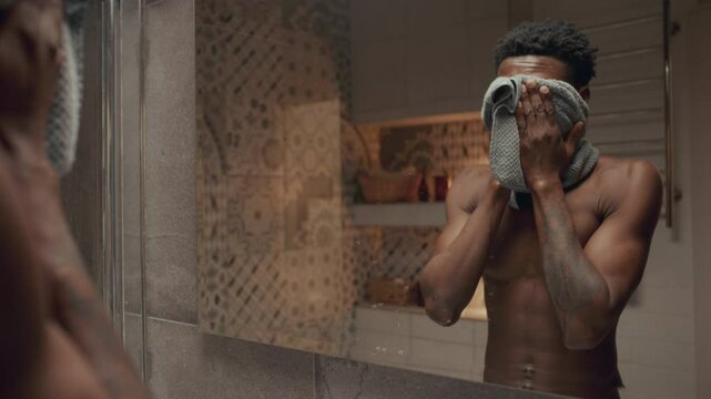 Young shirtless Black man washing his face with clean water, dru=ying skin with towel, looking in mirror and leaving bathroom