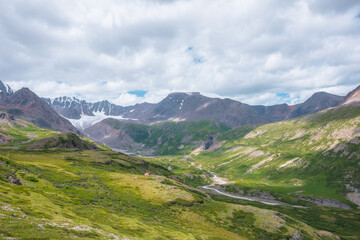 Wide green alpine valley with snake river among hills and rocks with vast view to large snow mountain top and big glacier tongue under cloudy sky. Serpentine river in high mountains. Shadows of clouds