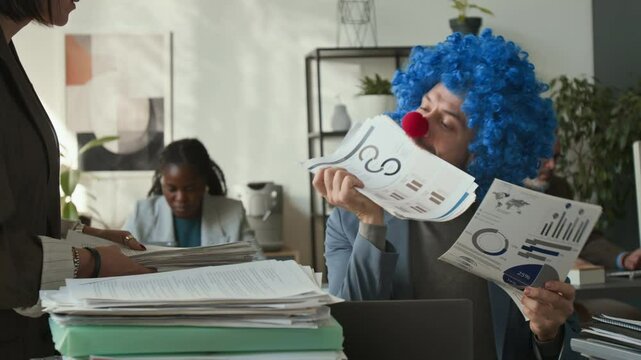 Office worker wearing clown wig and nose sitting at desk, holding documents with his teeth while receiving pile of papers from colleague, then typing on laptop with angry look