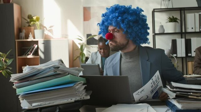 Funny businessman wearing clown wig and nose sitting at desk, receiving pile of papers from female colleague, making fist and using laptop