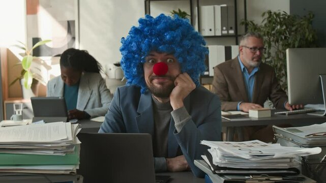 Portrait of businessman in formal suit, clown wig and nose typing on laptop at desk loaded with piles of papers, then looking at camera and sticking out his tongue during workday in office