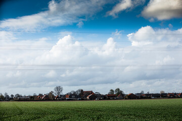 Rural Dutch landscape:  green field stretches to village with terracotta-roofed houses under a partly cloudy sky. Power lines cross the scene.