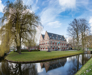 Canalside historical building, likely Dutch, with red shutters, reflected in calm water.  Springtime greenery surrounds the scene.