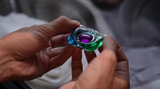 Close-up of colorful laundry detergent pod being held in hands, ready to be added to washing machine. Male putting liquid capsules with laundry detergent in washing machine tank, slow motion.