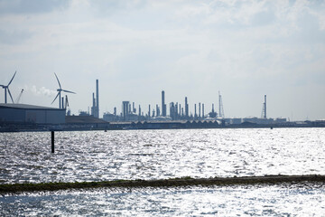 Obraz premium Industrial complex by a body of water, likely a harbor or river. Silhouetted structures and wind turbines are visible.