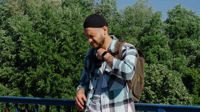Young bearded man stands on a bridge in Belgrade on a sunny spring day. A tourist guy with a backpack in a plaid shirt and a cap outdoor. Squints at the sun and puts on sunglasses