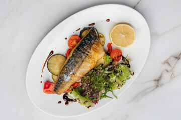 Grilled mackerel fillet is served on a white oval plate with grilled vegetables, fresh salad, lemon slice, and balsamic sauce on a marble table. Restaurant or cafe menu, cookbook recipe
