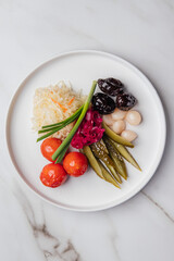 Displaying an assortment of pickled delicacies on a white plate, including sauerkraut, tomatoes, cucumbers, prunes, garlic, and red onions. Restaurant or cafe menu, cookbook recipe

