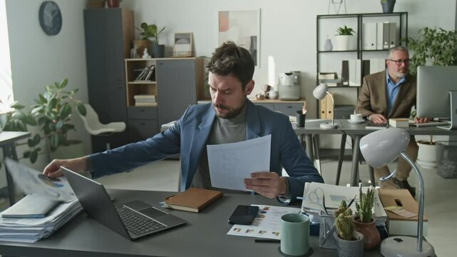 Concentrated businessman sitting at desk in open space office, reading documents and typing on laptop, then leaning back in chair and taking break during workday