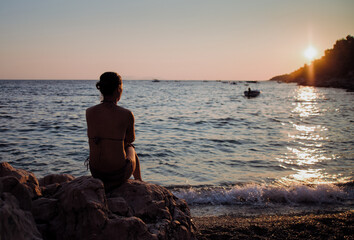 Woman sitting alone on rocks by the sea watching the sunset