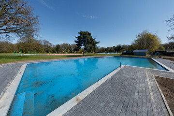 Fototapeta premium Under construction, a large blue pool sits amidst a park-like setting. Surrounding landscaping and playground equipment are visible.