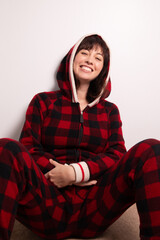Portrait of a caucasian woman wearing a red onesie pajamas for adults. She is sitting and leaning back and smiling. She is wearing the hood.  © Angela