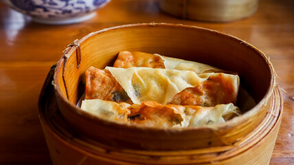 Three spring rolls or lumpia in a bamboo steamer basket, placed on a wooden table