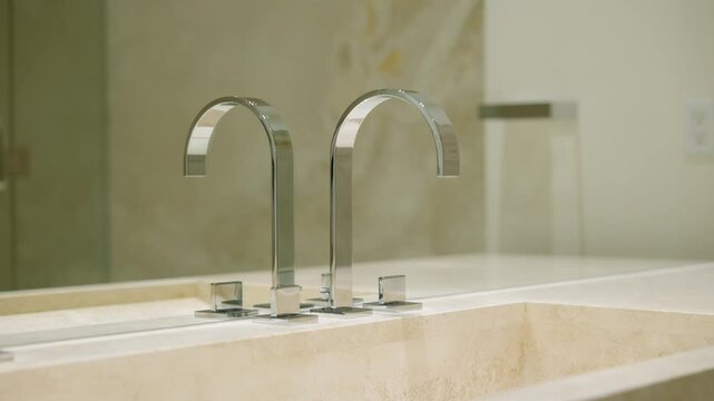 Modern bathroom sink with two sleek, chrome faucets. Light-colored stone material and mirror in background reflecting the faucets