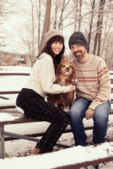Winter portrait of a caucasian couple sitting outside on a bench with their cocker spaniel dog. The couple are wearing warm sweaters, hats and pants. The trees behind them are bare and snow covered. 