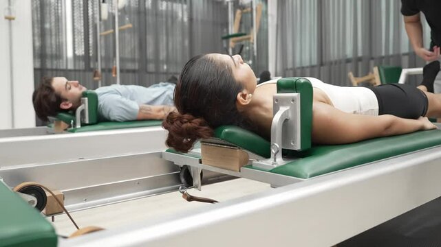 Couple practicing leg exercises on pilates reformer to strengthen core and improve flexibility at gym. Pilates partner doing pelvic bridge pose on reformer for improving lower back muscle. Habituate.