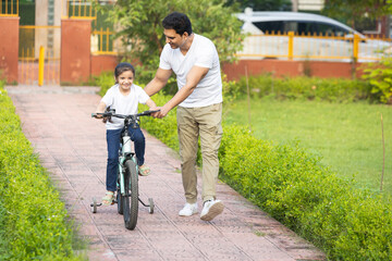 Young indian father teaching her daughter to ride bicycle in summer park outdoors. Dad helping and supporting her to cycle and pedal a bike.