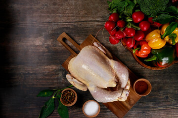 Whole chicken placed on wooden cutting board surrounded by vibrant vegetables. Fresh bell peppers, tomatoes, and greens create pleasing display for meal preparation.