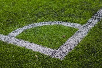 close-up view of a section of a sports field, specifically a corner area. The field is covered in green artificial turf with white painted lines marking the boundaries.