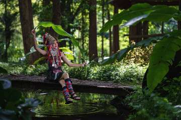 A young woman in traditional hill tribe attire is seated on a wooden bridge, holding a banana leaf in a lush green forest.