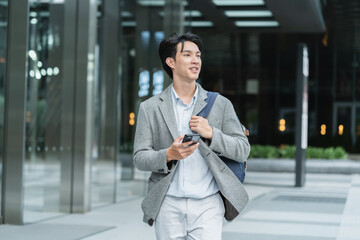 Portrait of young Asian man standing outdoor happy smiling use mobilephone.Handsome young asian man dressed casually walking spending time outdoors at city urban,carrying backpack,using mobile phone
