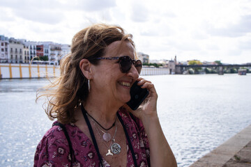 Smiling older woman with sunglasses answers a cell phone call on the city's river walk at sunset.