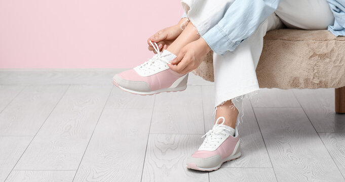Woman in stylish sneakers and jeans tying shoe lace against pink background