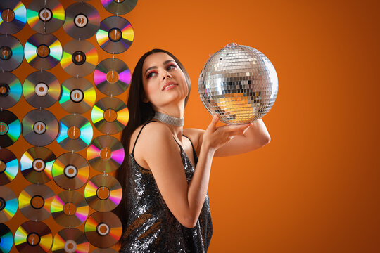 Stylish young woman with disco ball and compact disks on orange background