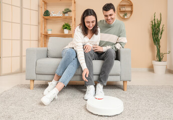 Modern robot vacuum cleaner cleaning carpet near relaxing couple on sofa