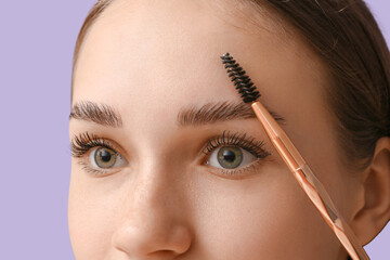 Young woman correcting shape of her eyebrows on lilac background, closeup