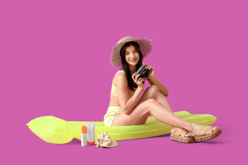 Young pretty woman in hat and swimsuit sitting on inflatable mattress with photo camera on purple background