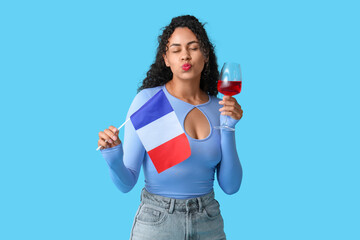 Beautiful young African-American woman with flag of France and glass of red wine blowing kiss on blue background