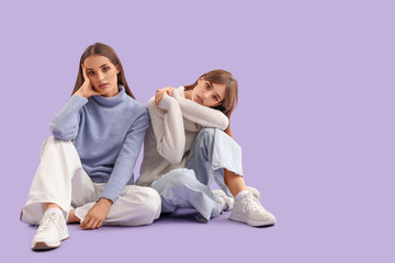 Beautiful young fashionable sisters in stylish outfits sitting on purple background