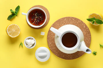 Stylish tea pot, cup of tea, lemon and green leaves on yellow background