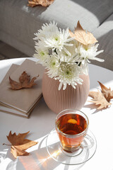 Cup of hot tea, books, autumn leaves and vase with flowers on coffee table in living room, closeup
