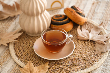 Wicker mat with cup of hot tea, tasty poppy buns and autumn leaves on table at home, closeup