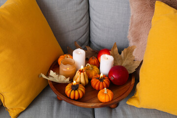 Wooden tray with glowing candles, apples and autumn leaves on sofa in room