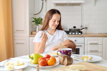 A mother smiles as she feeds her baby while seated at a table filled with breakfast foods in a bright, warm kitchen. Fresh fruits and bread are neatly arranged