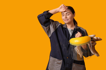 Tired female construction worker with protective helmet and gloves on yellow background. Woman in...