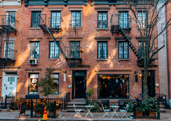 A cozy brick building with fire escapes, storefronts, and outdoor seating in a quiet New York City street, capturing urban charm and character...