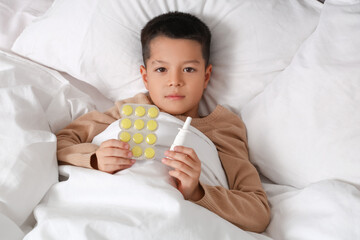 Ill little Asian boy with pills for sore throat and nasal drops lying in bed, top view