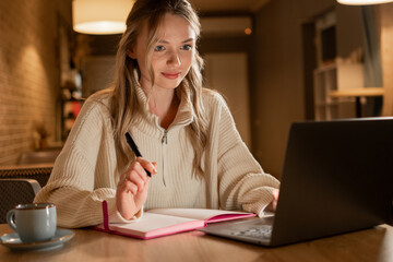 Concept of Distance Education. Young female student learning online in the cafe, looking at laptop, writing notes in diary