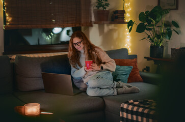 Obraz premium A young woman sitting comfortably on a couch using a laptop during a cozy and festive evening at home