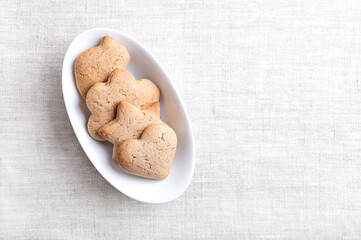 Homemade gingerbread cookies in an oval white bowl on linen with empty space for text. Sweet, light brown and flat baked goods for Christmas season, flavored with ginger, cloves, nutmeg, and cinnamon.