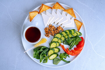 Healthy Breakfast Plate with Tea, Cheese, Fresh Vegetables and Toast, Top view of a healthy breakfast plate featuring tea, sliced cheese, fresh vegetables, toast, and walnuts on a white plate.